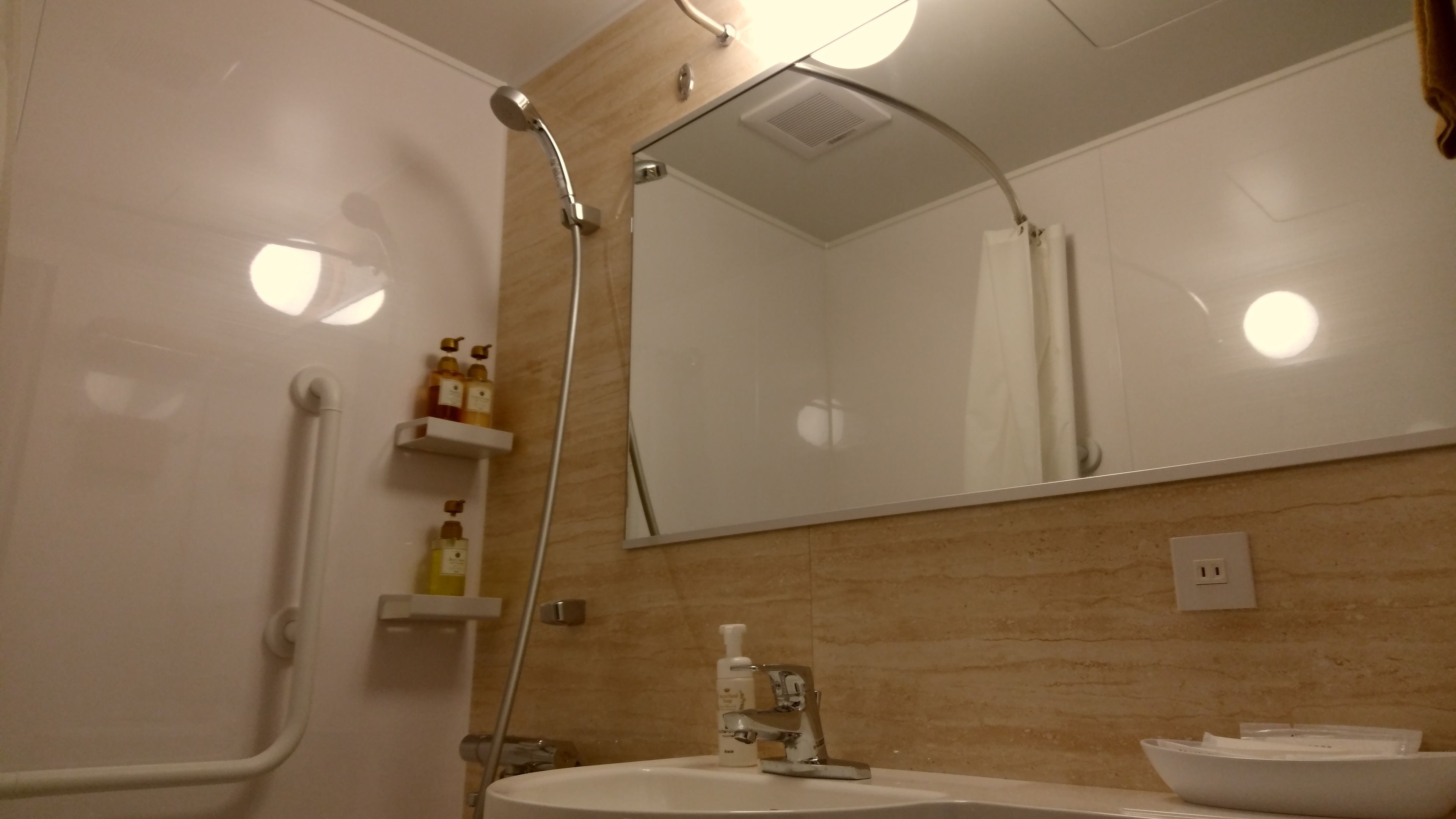 Casa de banho com lavatório branco, espelho, chuveiro com chuveiro manual e prateleira com artigos de higiene pessoal.