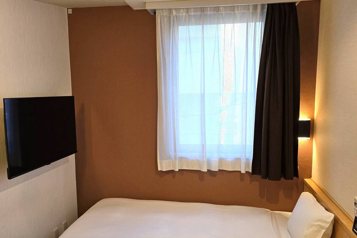 A hotel room with a bed, a flat-screen TV mounted on the wall, a window with sheer curtains, and a wall-mounted light.