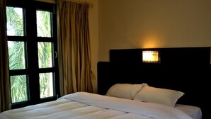 A neatly made bed with white linens, a headboard with a light fixture, and a window with curtains. - Rhino Lodge & Hotel (Sauraha)