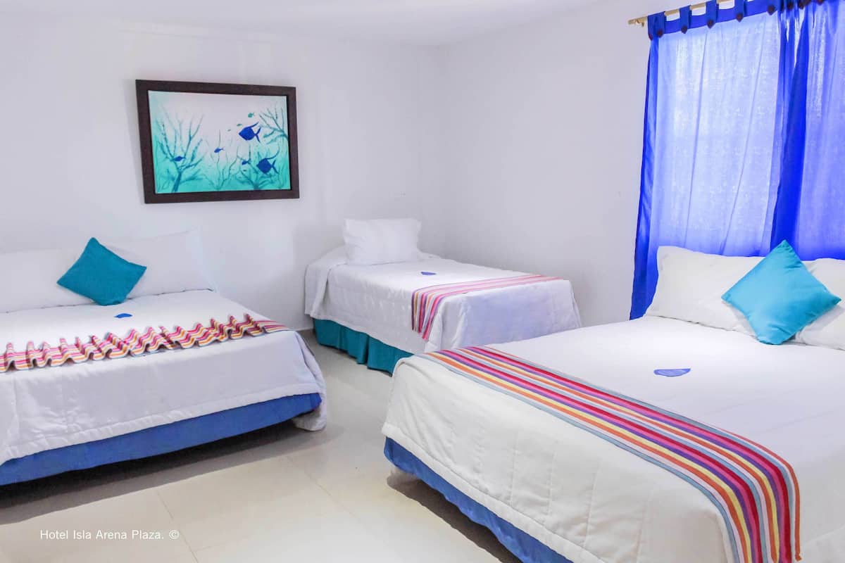 Une chambre d’hôtel avec trois lits, chacun recouvert d’une courtepointe blanche agrémentée de touches de bleu. Un tableau encadré orne le mur et un rideau bleu est accroché à la fenêtre.