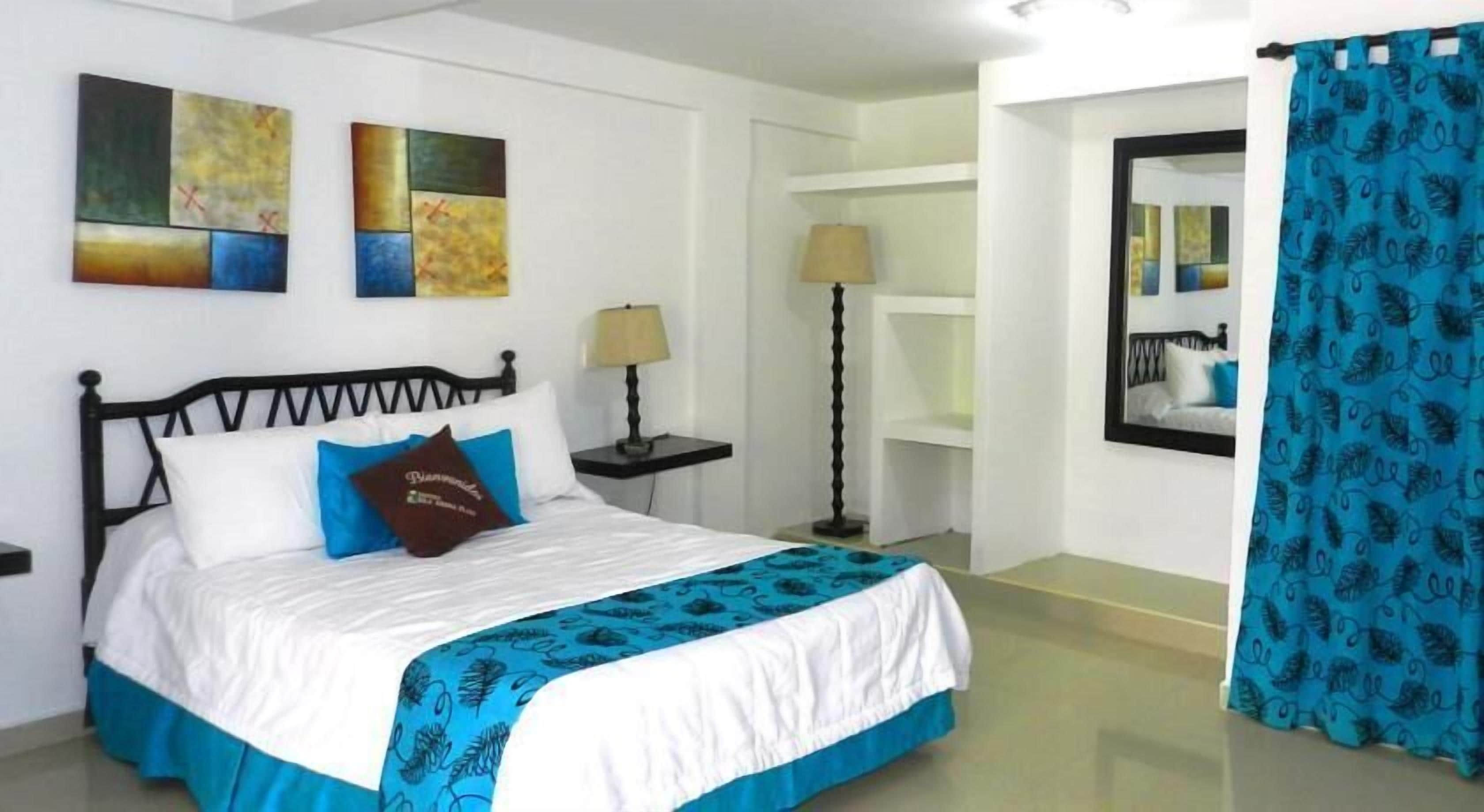 Une chambre à coucher avec un lit, une table de chevet, une lampe, un mur orné d’œuvres d’art abstraites, un miroir et un rideau bleu.