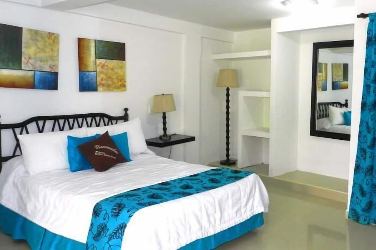 Une chambre à coucher avec un lit, une table de chevet, une lampe, un mur orné d’œuvres d’art abstraites, un miroir et un rideau bleu.