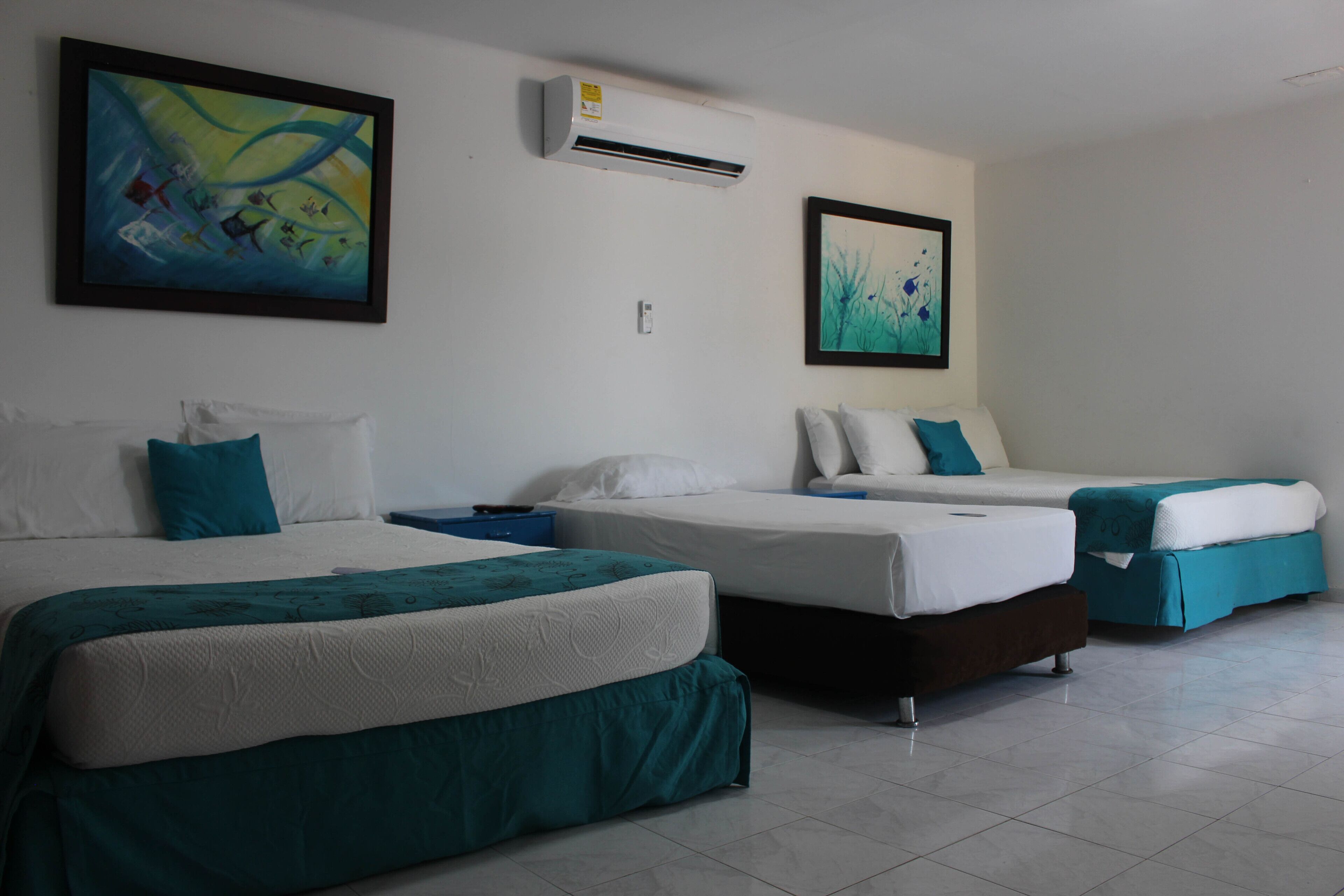 Habitación de hotel con tres camas, cada una con cubrecama verde azulado y blanco, y dos cuadros en las paredes.