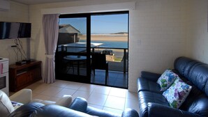 A living room with a black leather sofa, a television on the left, and a view of the beach through a glass door. - Marcel Towers Holiday Apartments (Nambucca Heads)