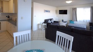 A modern living room with a grey sofa, a white dining table, and a kitchen area with cabinets and a countertop. - Marcel Towers Holiday Apartments (Nambucca Heads)
