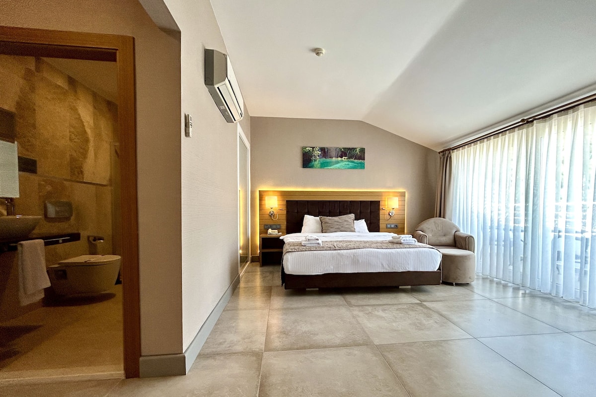 A modern hotel room with a large bed, a sitting area, a bathroom, and a view through large windows.