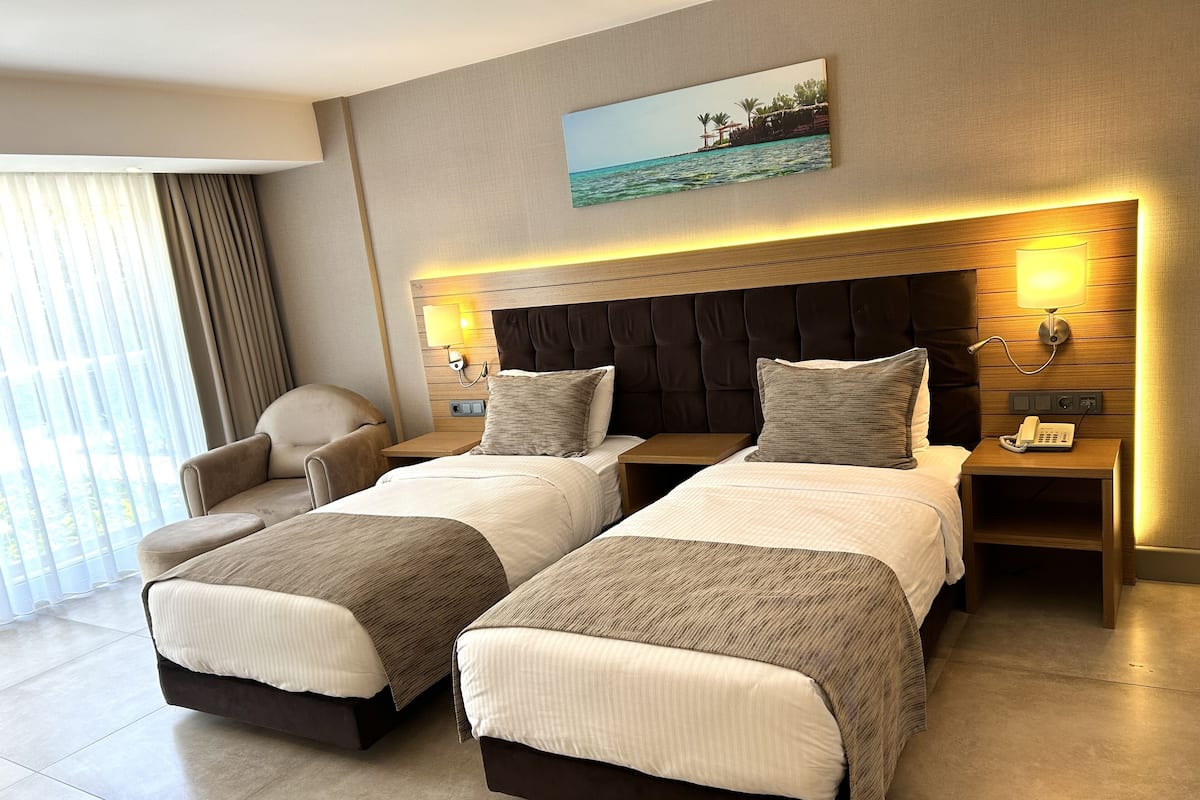 A hotel room with two beds, a chair, a painting, and a view of the sea.