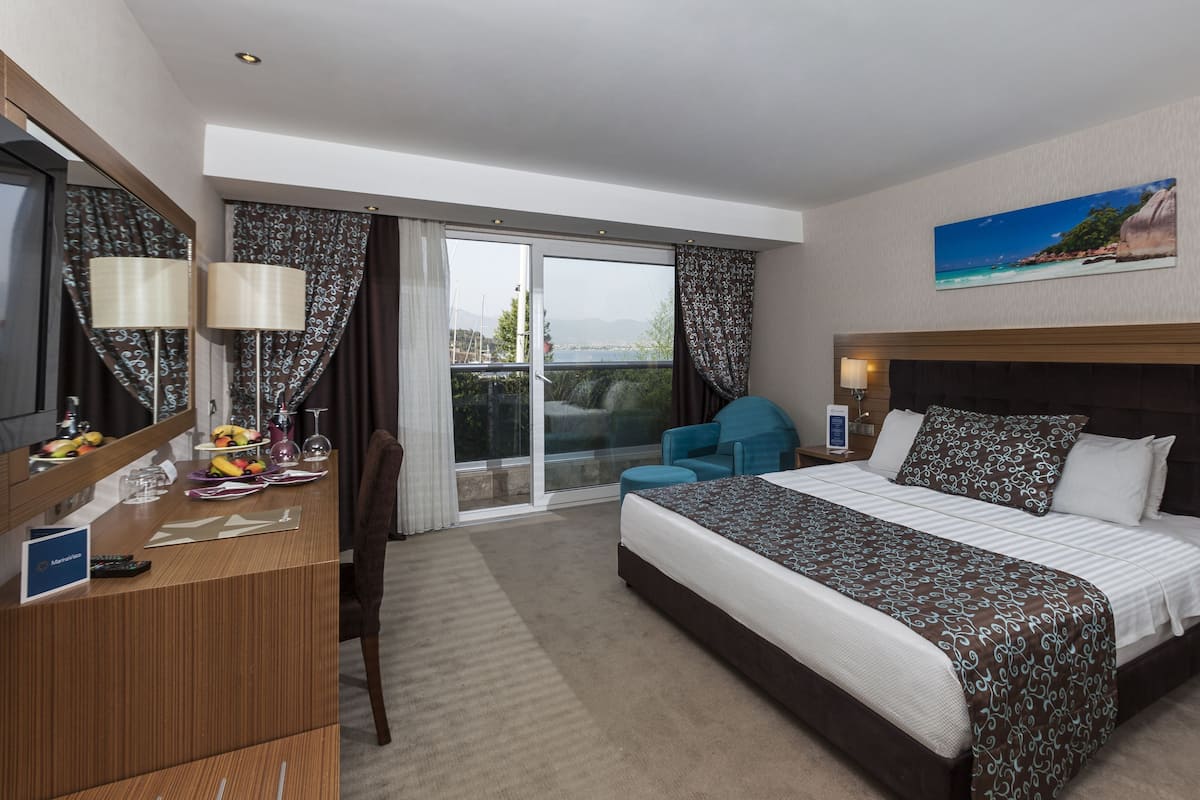 A hotel room with a bed, a desk with a computer, a chair, a desk lamp, a nightstand, and a painting on the wall.