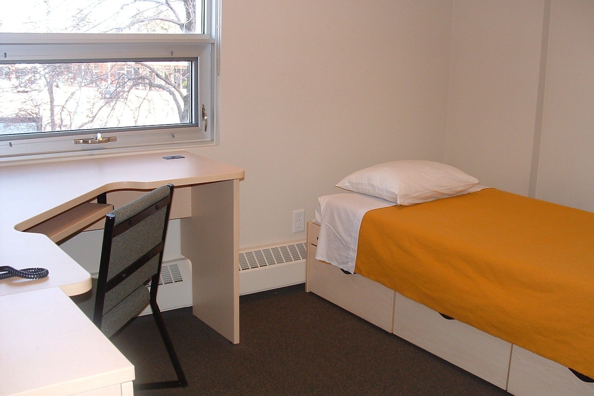 Une chambre de dortoir équipée d’un bureau, d’une chaise, d’un lit et d’une fenêtre.