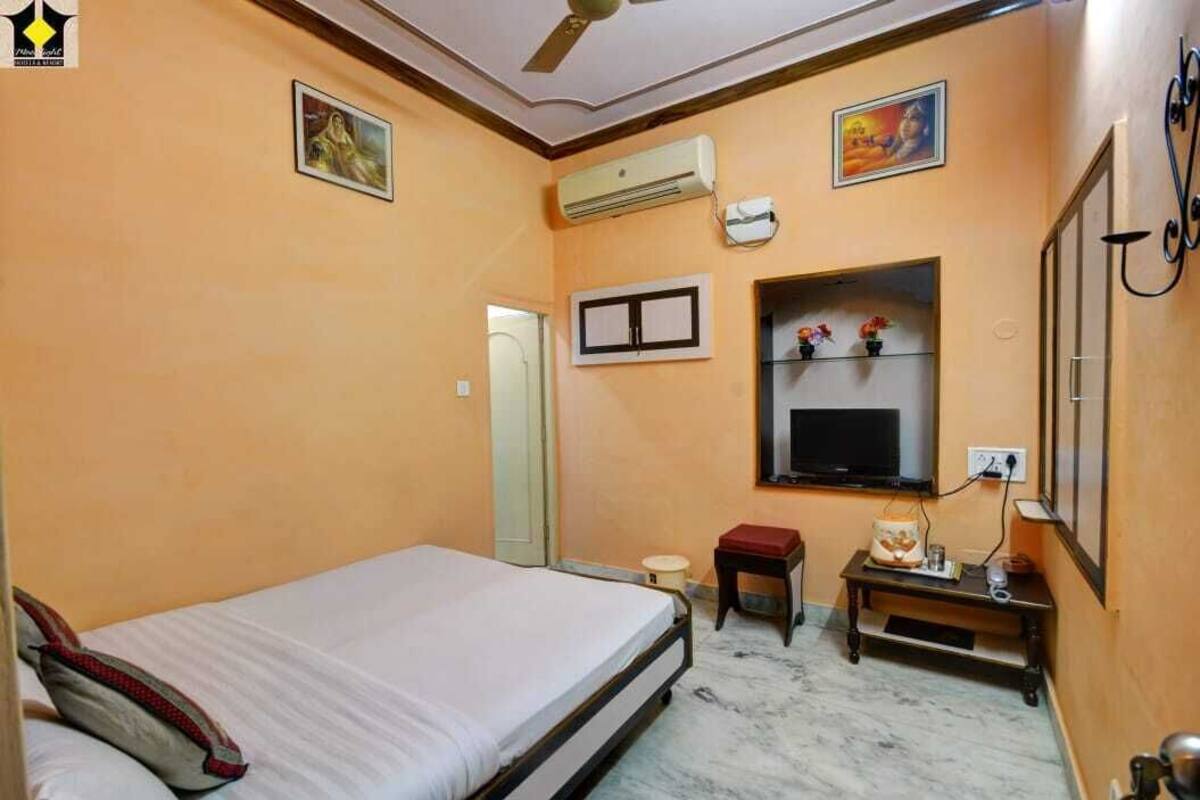 Een slaapkamer met een bed, een aan de muur bevestigde televisie, een airconditioning en een klein tafeltje met een lamp.