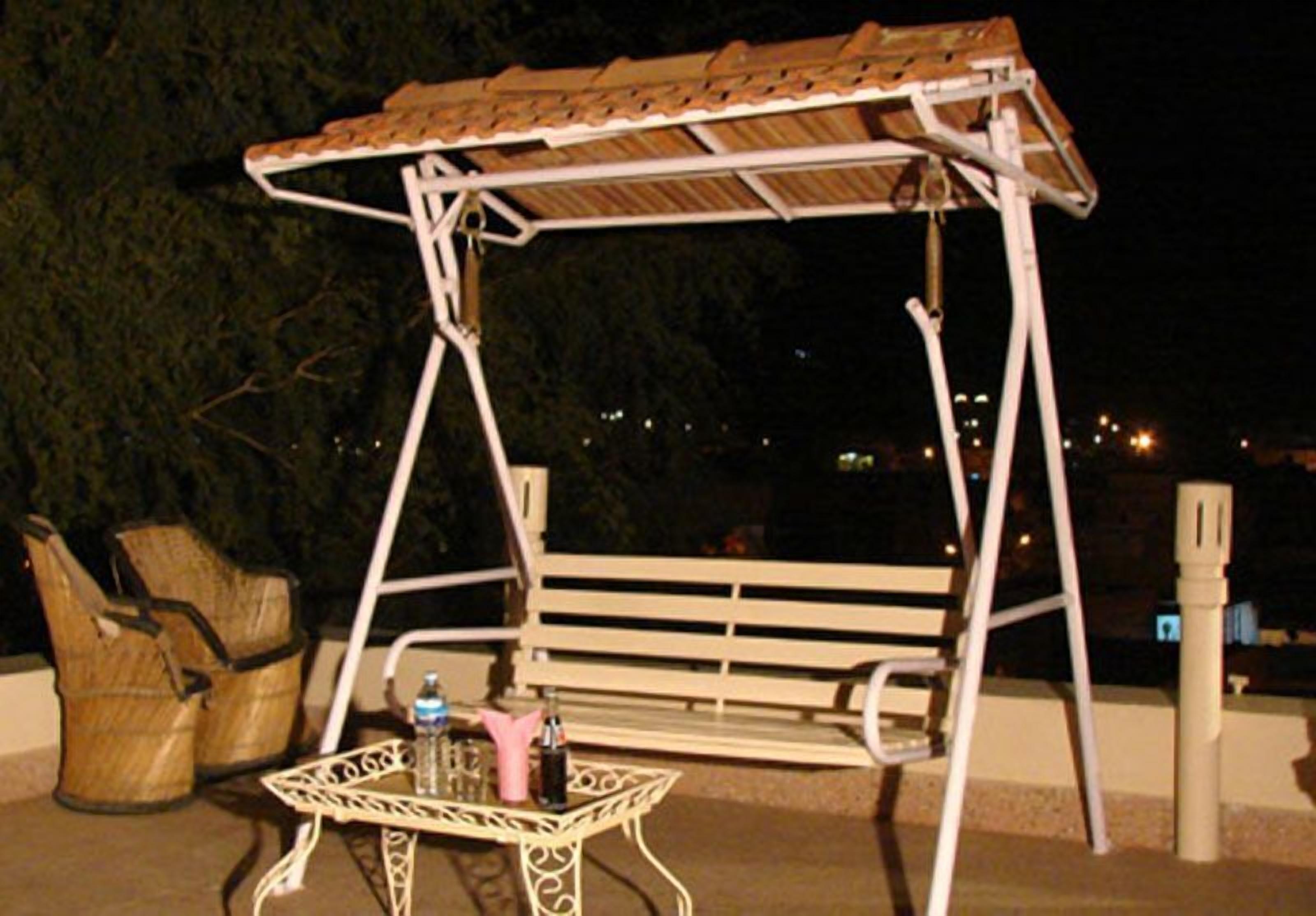 A white swing set with a wooden roof, a metal table with a bottle and a glass, and a wicker chair.