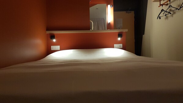Una habitación de hotel con cama, un espejo encima y una lámpara fijada a la pared.