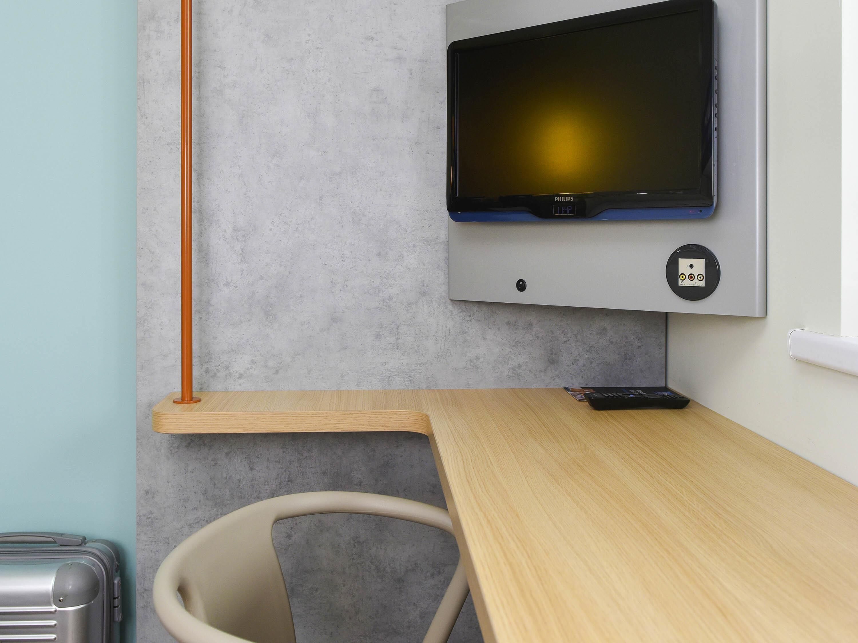 A modern room with a flat-screen TV mounted on the wall, a wooden desk, a chair, and a suitcase.