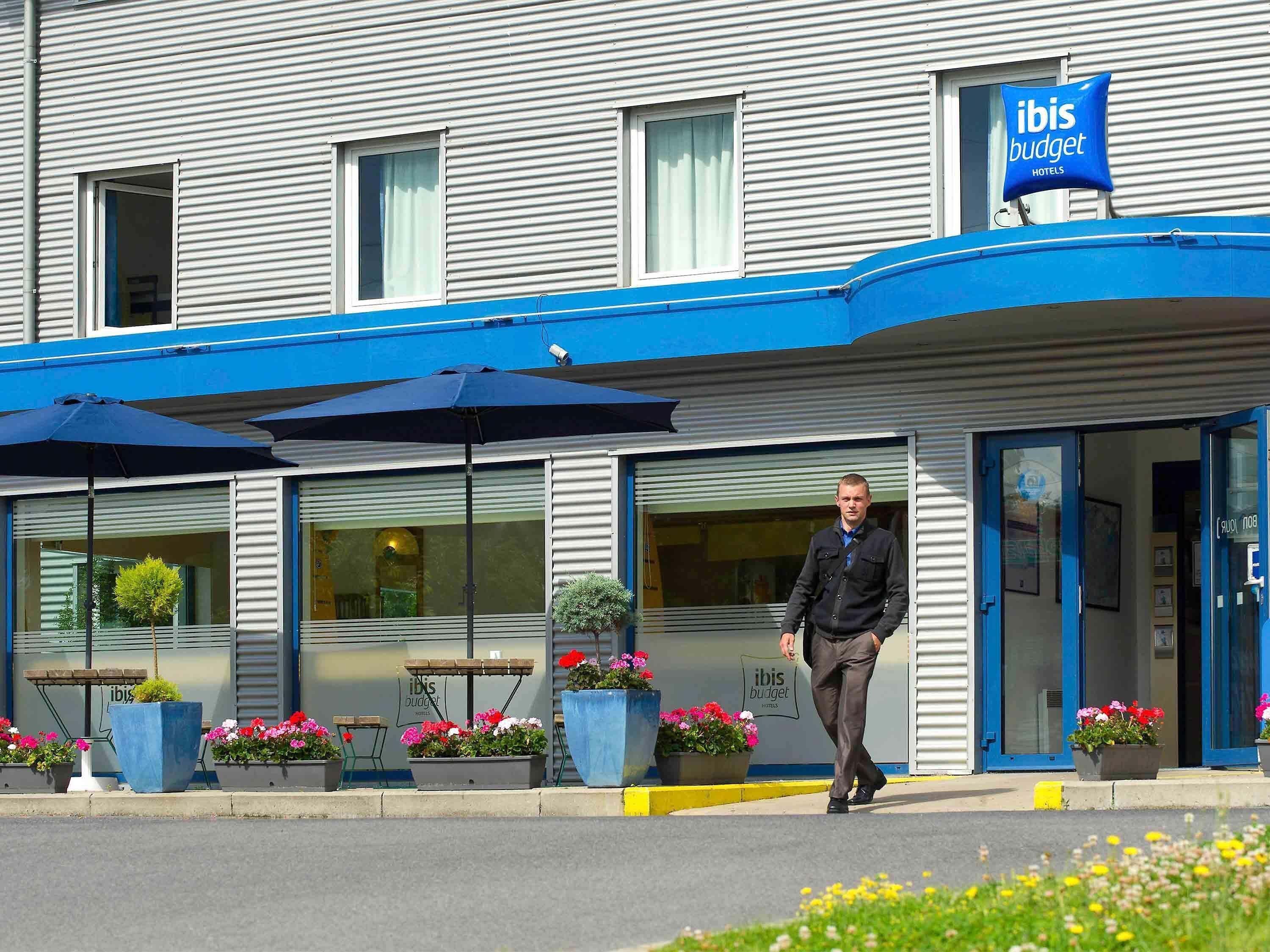 L’entrée d’un hôtel, avec un auvent bleu, des plantes en pot et une enseigne indiquant qu’il s’agit d’un hôtel ibis budget.