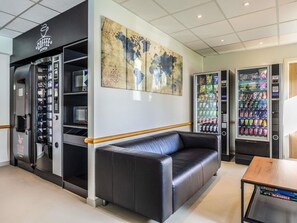 A modern interior with a coffee machine, vending machines, a leather couch, and a coffee shop sign. - Ibis budget Charleroi Airport (Fleurus)