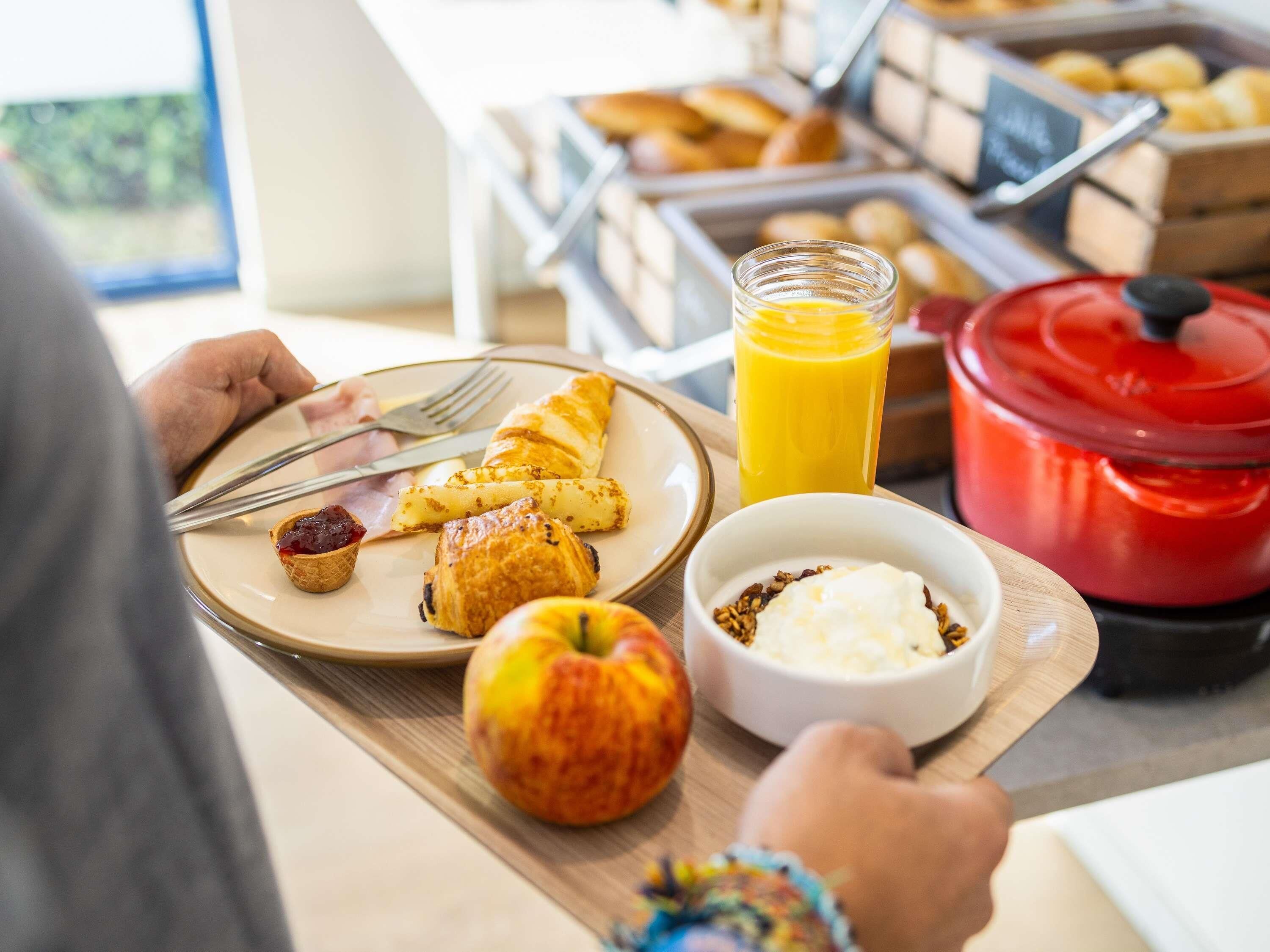 Una bandeja de desayuno con croissants, un muffin, una manzana, un vaso de zumo de naranja y un cuenco de cereales.