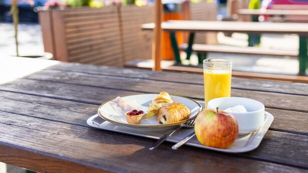 Puinen piknikpöytä, jonka päällä on lautanen täynnä ruokaa: croissant, omena ja lasi appelsiinimehua.