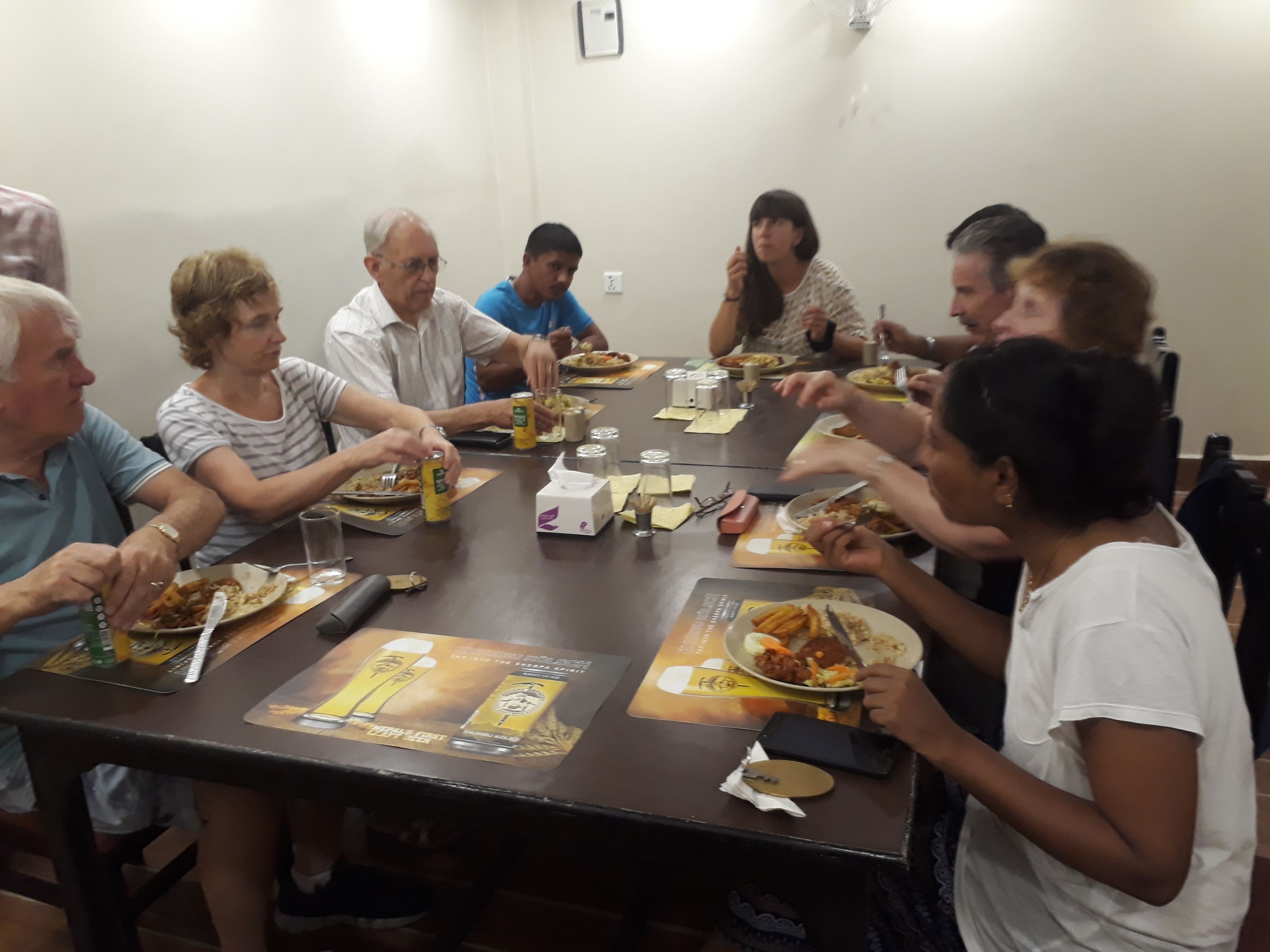 A group of people seated around a rectangular table with a brown tablecloth, plates of food, and a few cans of beer.