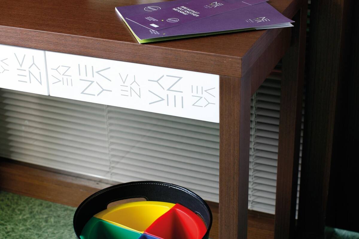 Un bureau avec un écran d’ordinateur, une petite plante et quelques papiers. On y voit une poubelle avec des compartiments de couleurs vives et, au fond, une fenêtre avec des stores.