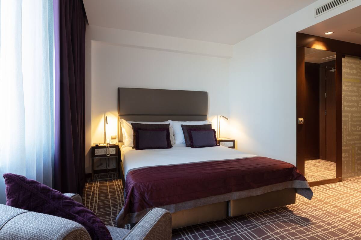 Une chambre d’hôtel avec un grand lit, deux tables de chevet, une petite table, une chaise et une vue sur l’extérieur par la fenêtre.