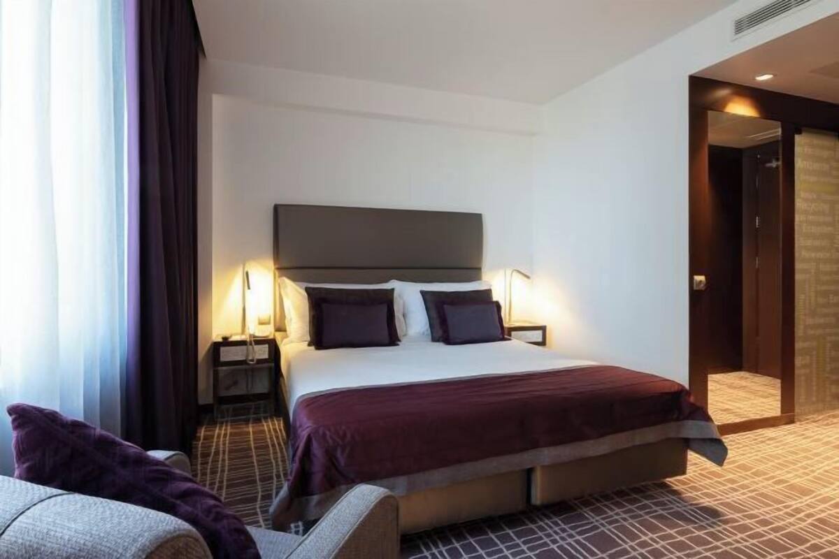 Une chambre d’hôtel avec un grand lit, des lampes de chevet, un fauteuil et une vue sur le couloir.