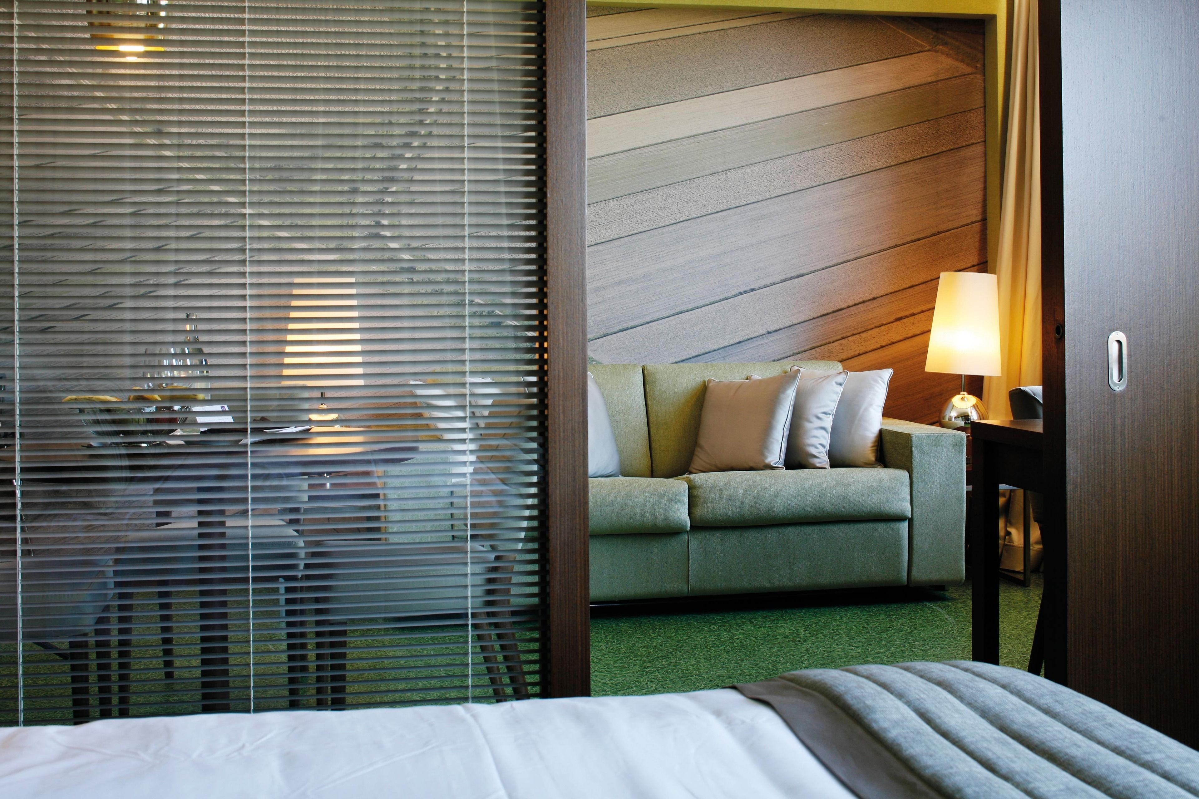 Habitación de hotel con cama, sofá verde, lámpara y pared de madera.