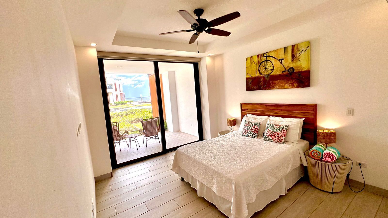A bedroom with a bed, a ceiling fan, a painting, a nightstand, and a view through a glass door to an outdoor area with chairs.