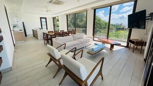 A spacious living area with a sofa, chairs, a coffee table, and a TV. There is a balcony with a view of greenery.