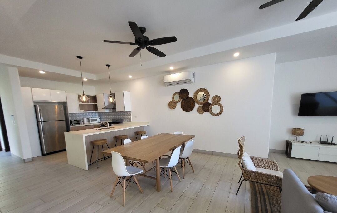 Cozinha moderna com ilha central, armários brancos e eletrodomésticos de aço inoxidável. Possui um espaço de refeições com mesa de madeira e cadeiras brancas.