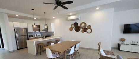 Cozinha moderna com ilha central, armários brancos e eletrodomésticos de aço inoxidável. Possui um espaço de refeições com mesa de madeira e cadeiras brancas.