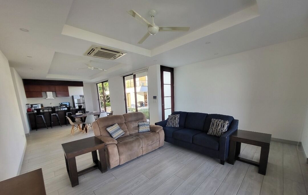 A spacious living area with a sofa, a coffee table, and a dark-colored couch. There is a kitchen in the background with modern appliances and a dining area.