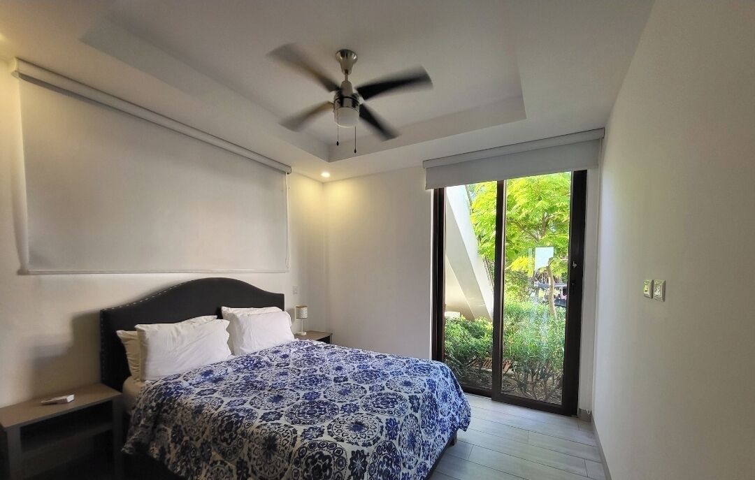A bedroom with a bed, a ceiling fan, a nightstand, and a view of greenery through a sliding glass door.