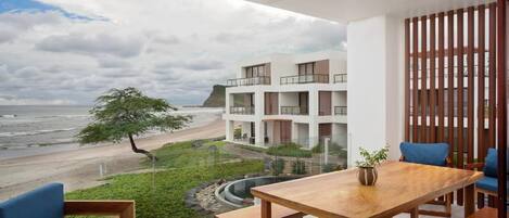 A modern balcony with a wooden table and blue chairs, overlooking a beach and a building with multiple balconies.