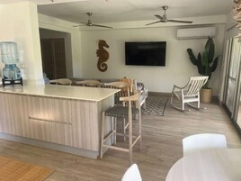 Una cocina moderna con una isla central, taburetes de bar, ventiladores de techo, un televisor montado y una planta en maceta.