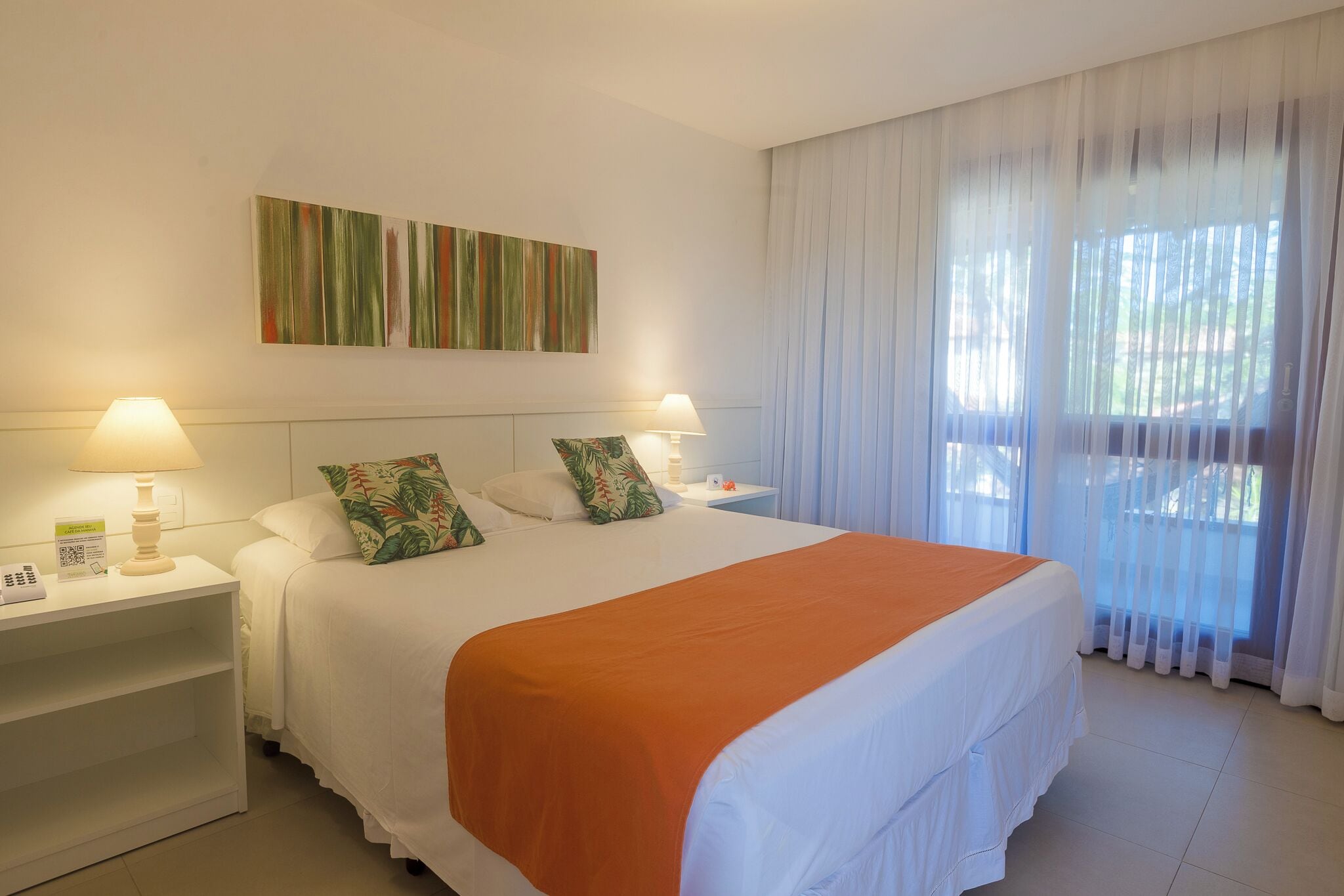 A hotel room with a large bed, two bedside tables, a nightstand, a window with sheer curtains, and a wall-mounted artwork.