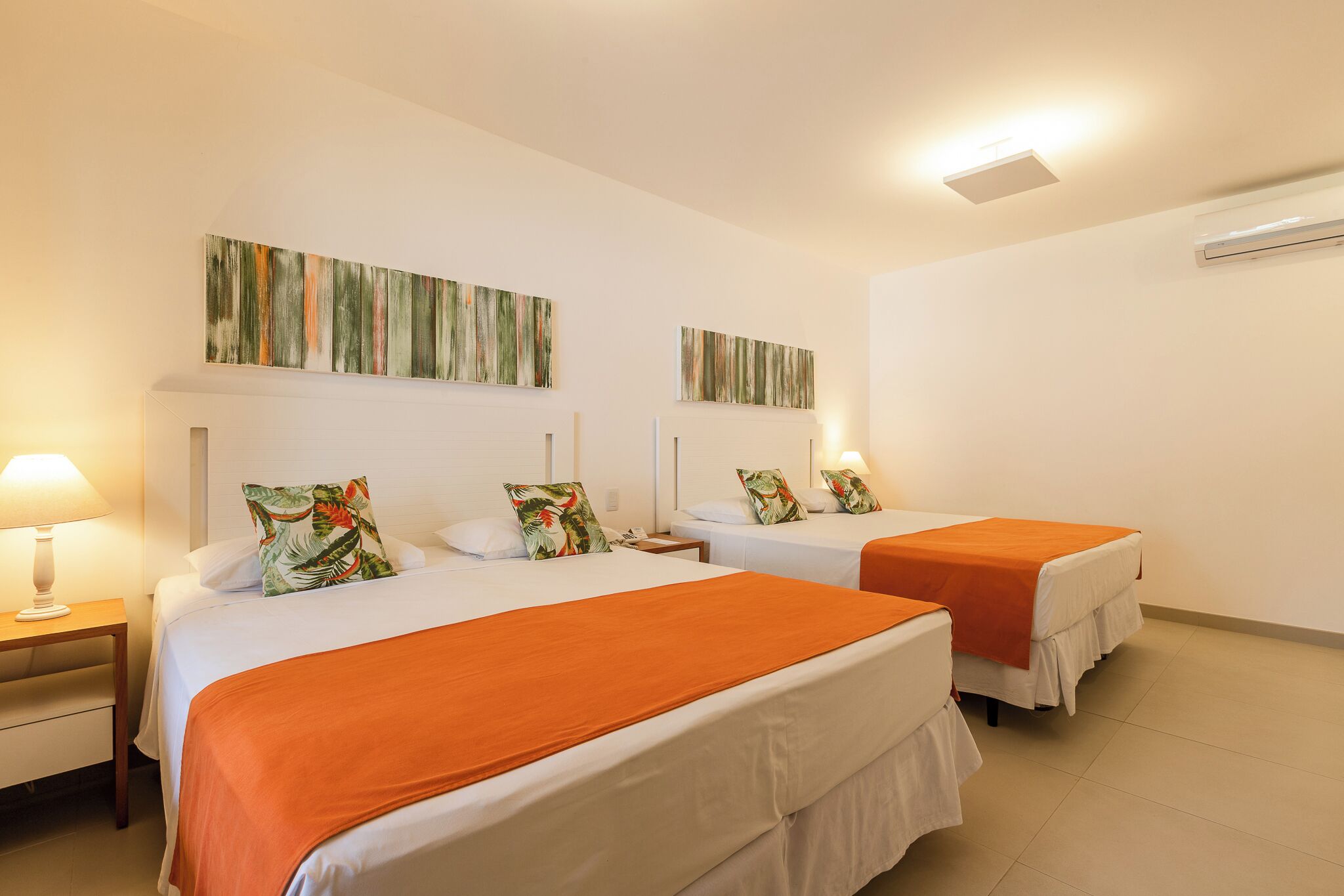 A hotel room with two beds, each with an orange bedspread, white bedding, and a tropical-themed pillow. There is a nightstand with a lamp, and the room has a white wall with two abstract paintings.