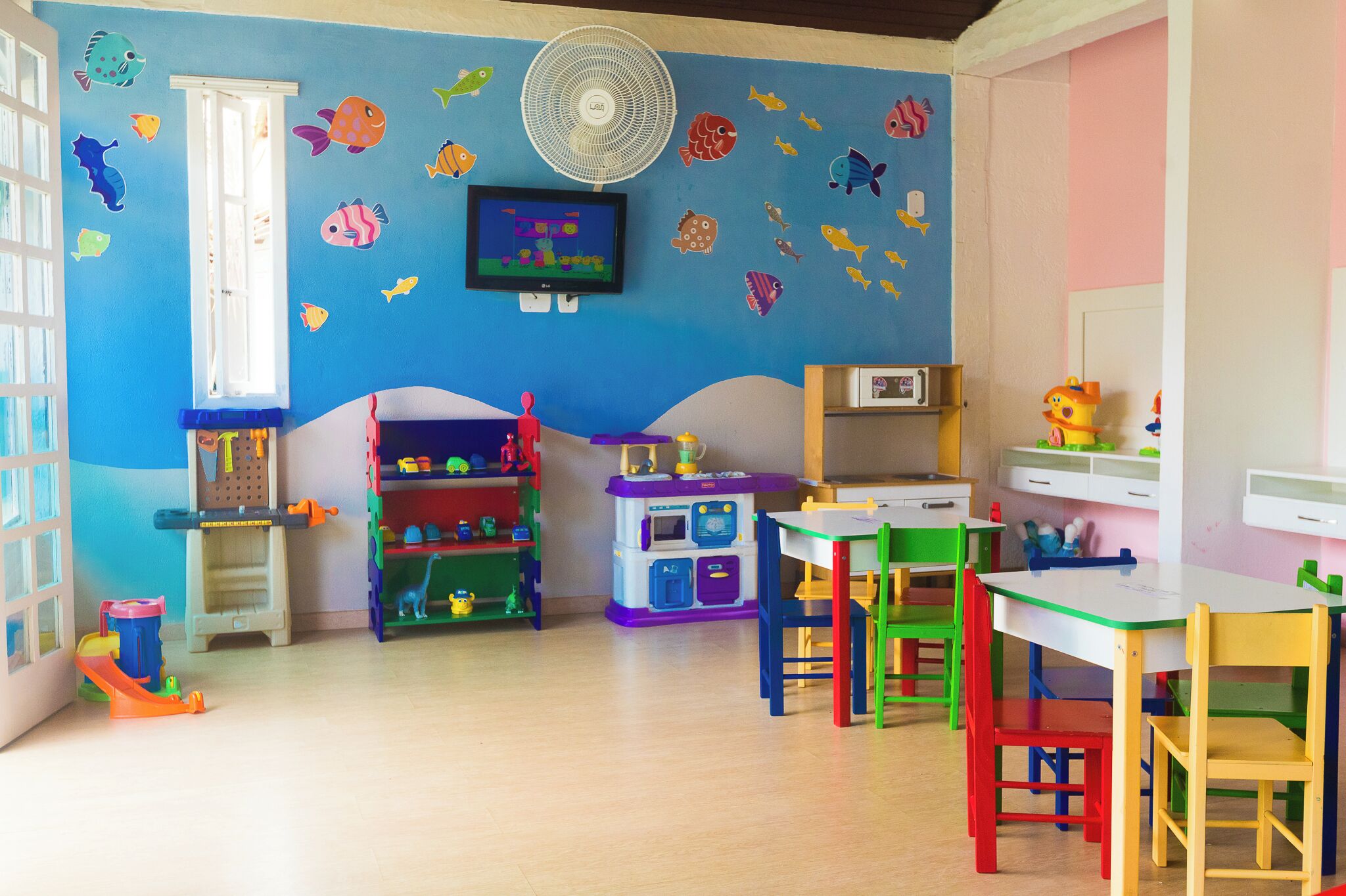 Une salle de jeux pour enfants avec un mur bleu, des autocollants représentant des poissons colorés, un ventilateur et divers meubles pour jouer.