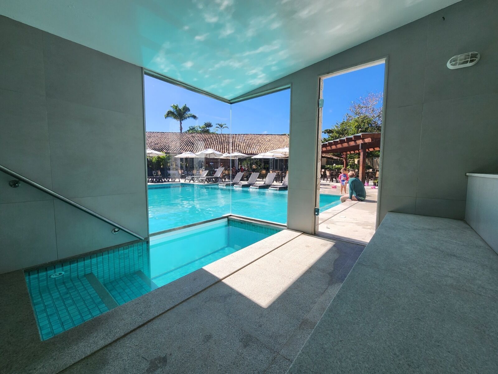 A modern indoor swimming pool with clear blue water, a glass ceiling, and a view of an outdoor pool area with umbrellas and lounge chairs.