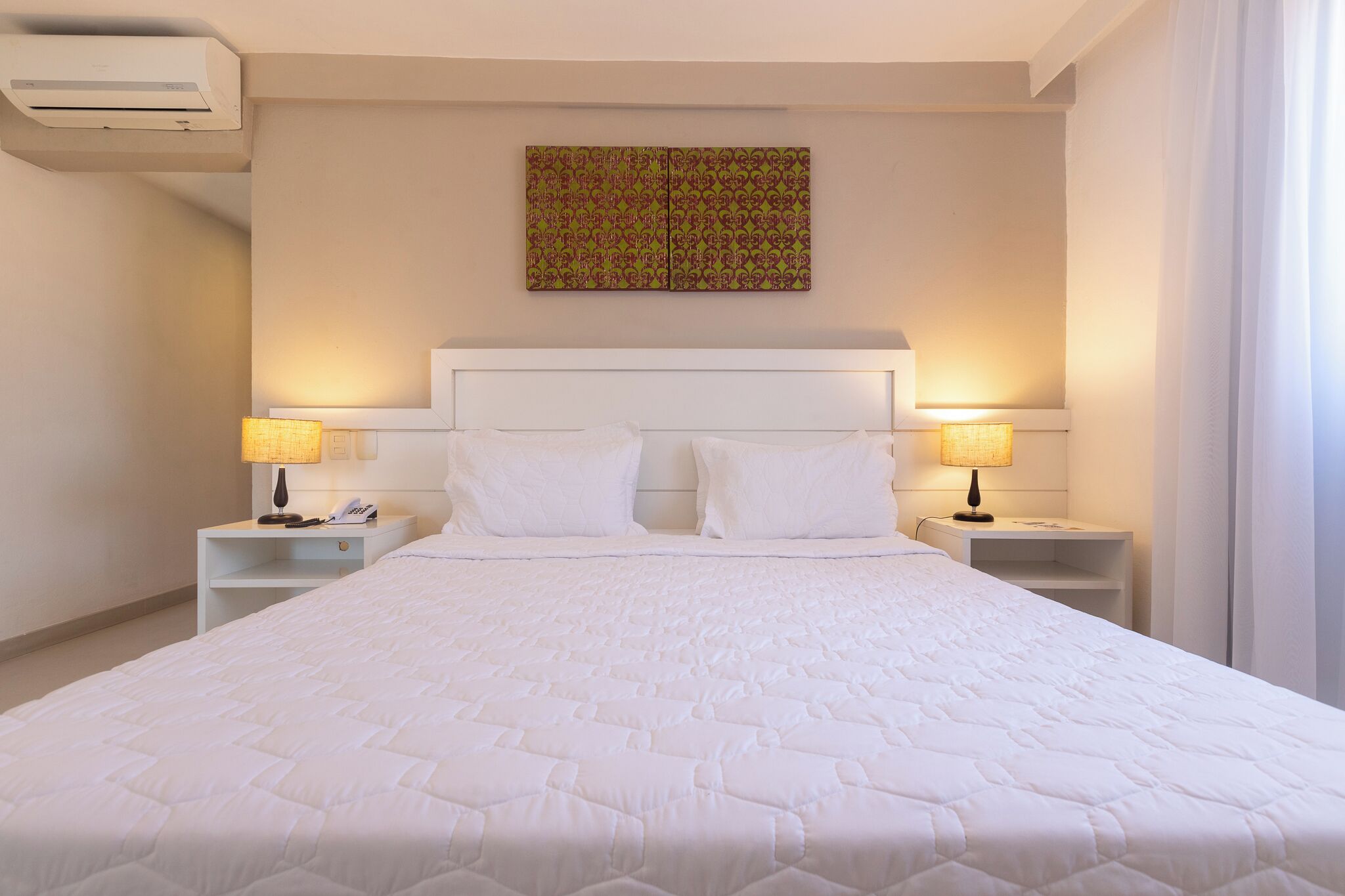A hotel room with a large bed, two bedside tables with lamps, a wall-mounted air conditioner, and a decorative wall piece.