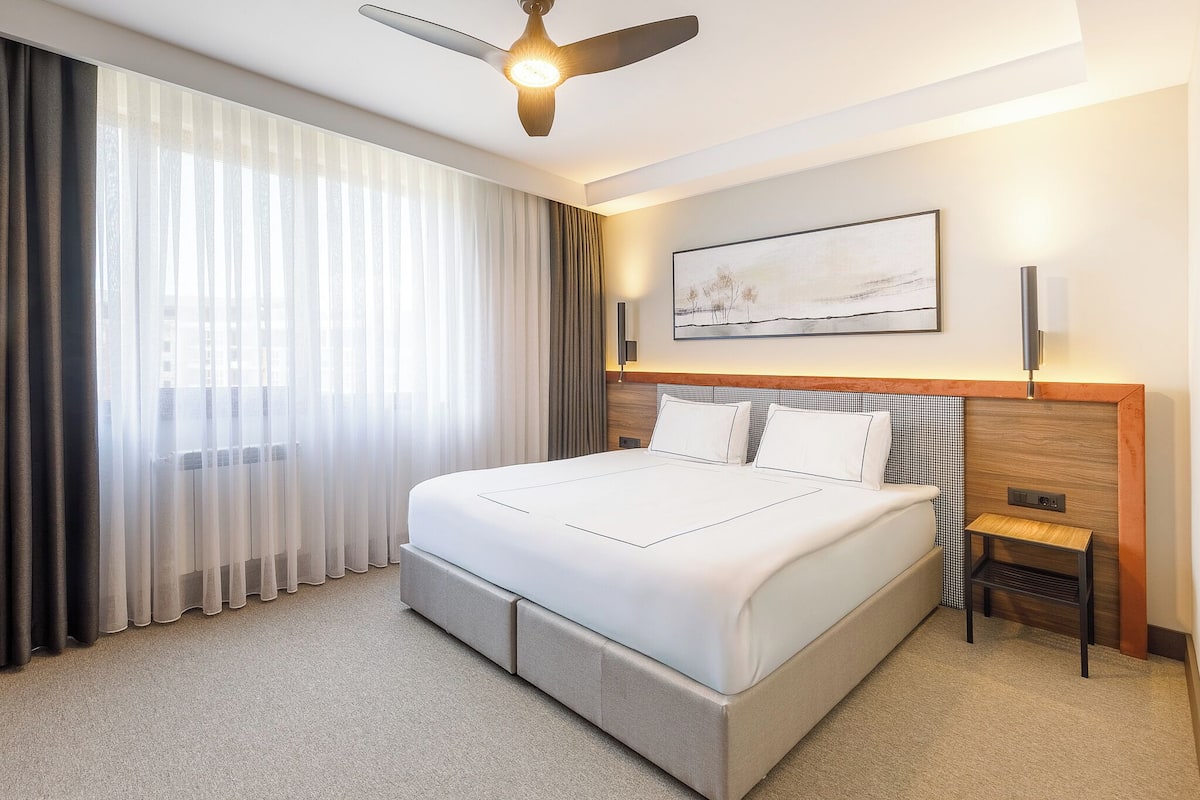 A modern hotel room with a large bed, a ceiling fan, a nightstand, and a painting on the wall.