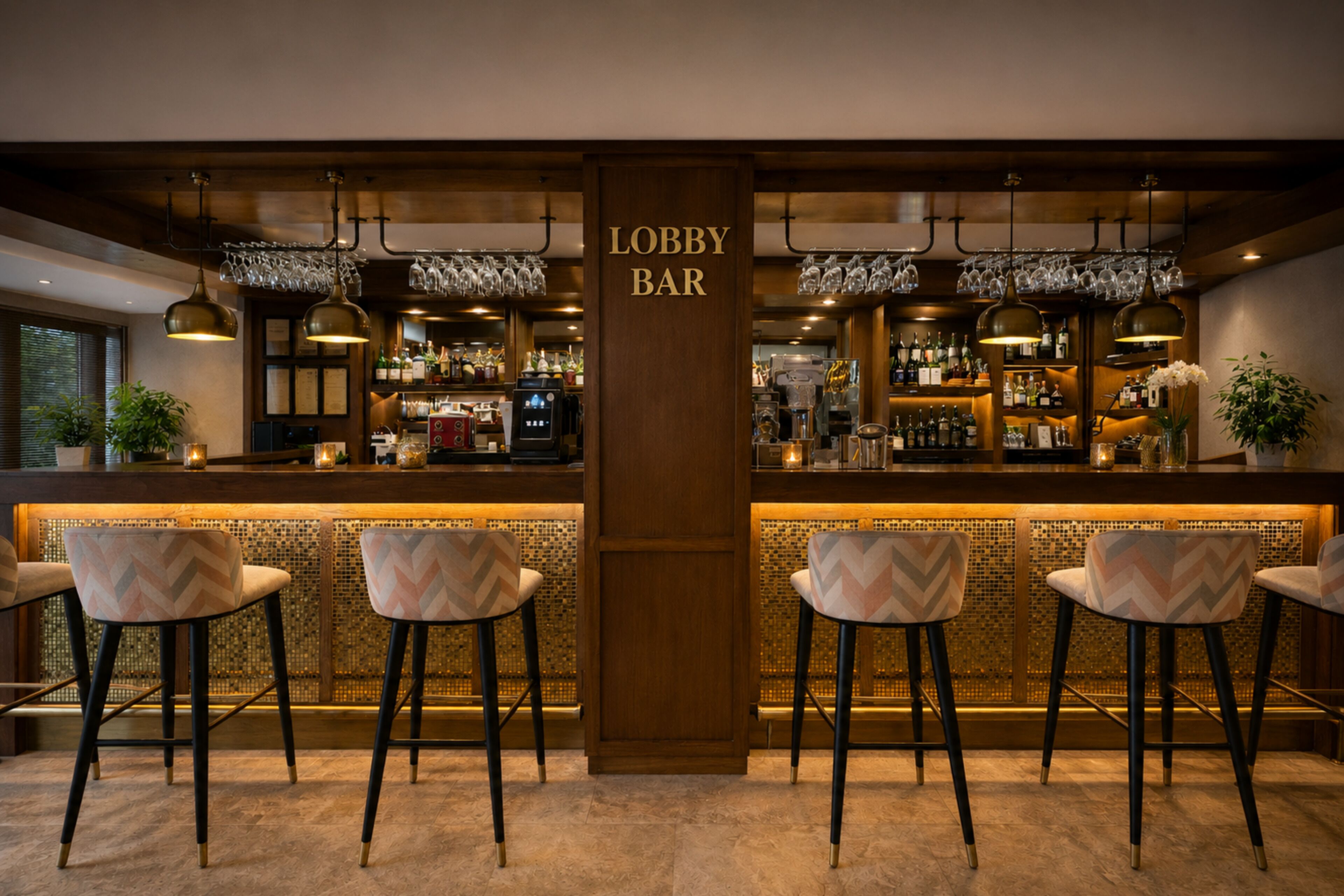 Angolo bar con un cartello che indica 'Lobby Bar', completo di sgabelli, un bancone con diverse bottiglie e bicchieri appesi.