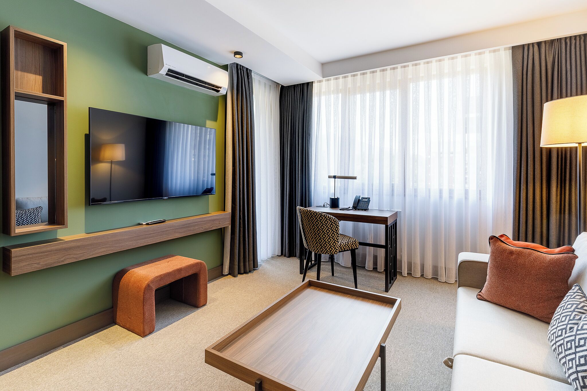 A modern hotel room with a flat-screen TV, a desk with a chair, a sofa with a patterned pillow, and a wooden bench.