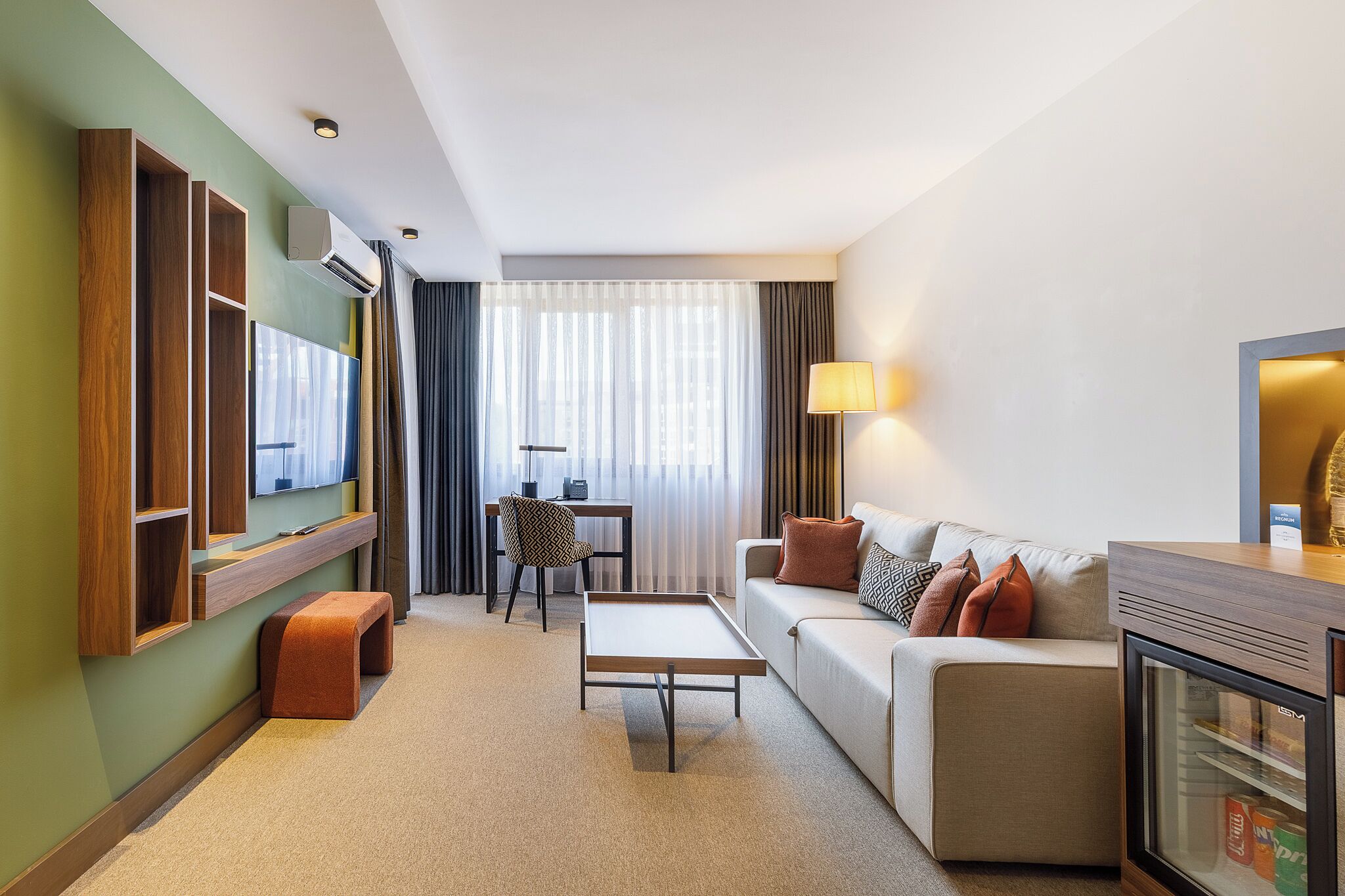 Una habitación de hotel moderna con sofá, una mesita, televisión, escritorio y mini refrigerador.