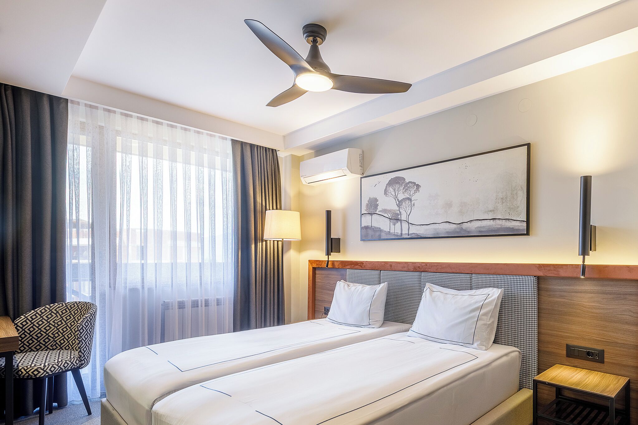 A hotel room with a large bed, a desk, a chair, a ceiling fan, and a painting on the wall.