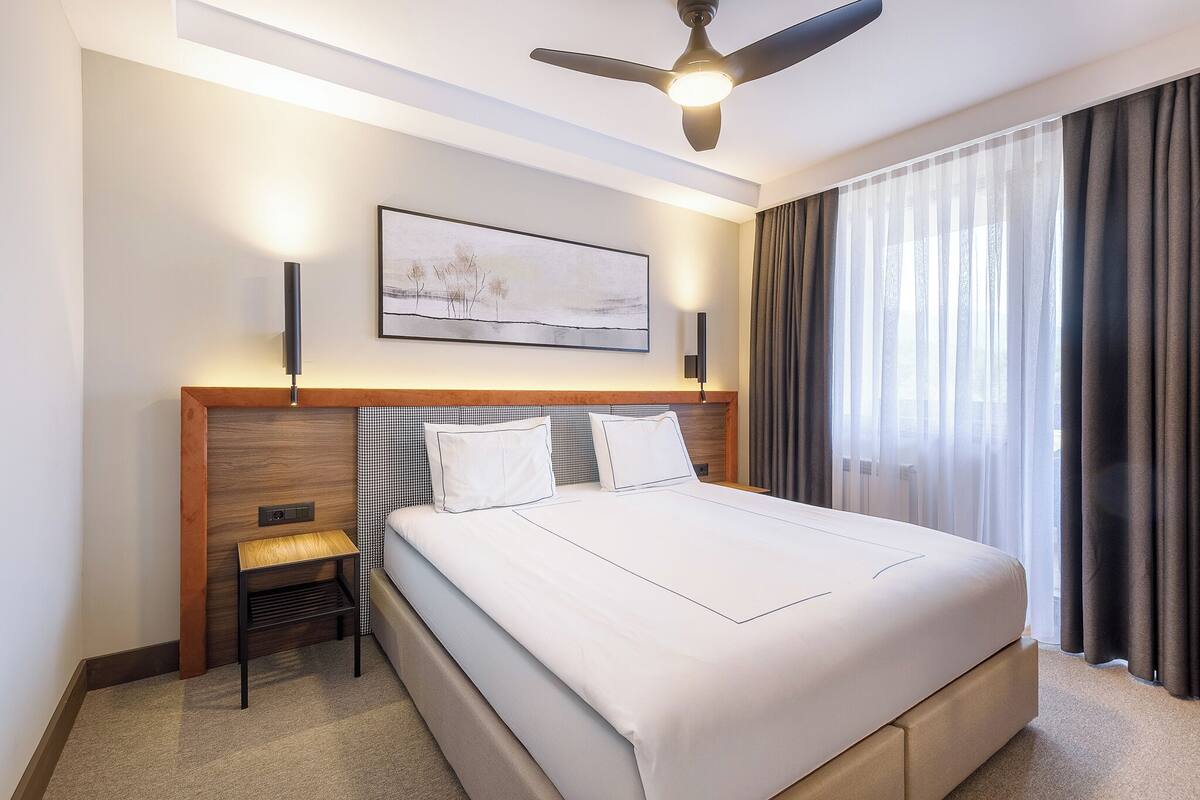 A hotel room with a large bed, a ceiling fan, a wall-mounted artwork, and a window with curtains.