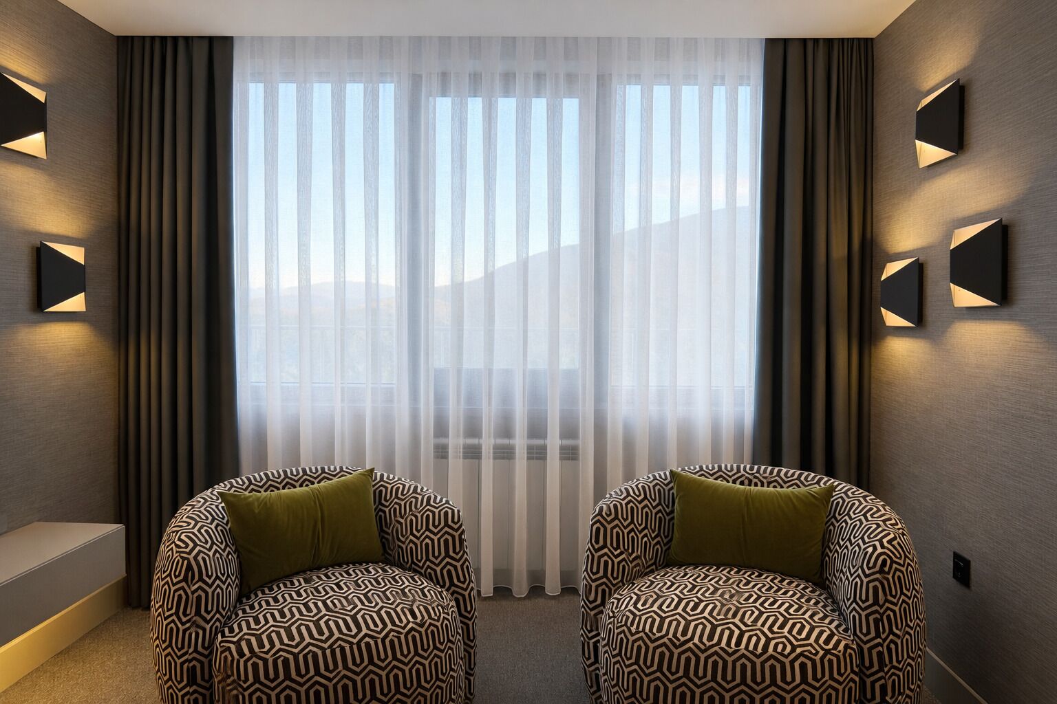 Una habitación moderna con dos sillones estampados, un ventanal con cortinas translúcidas e iluminación en la pared.
