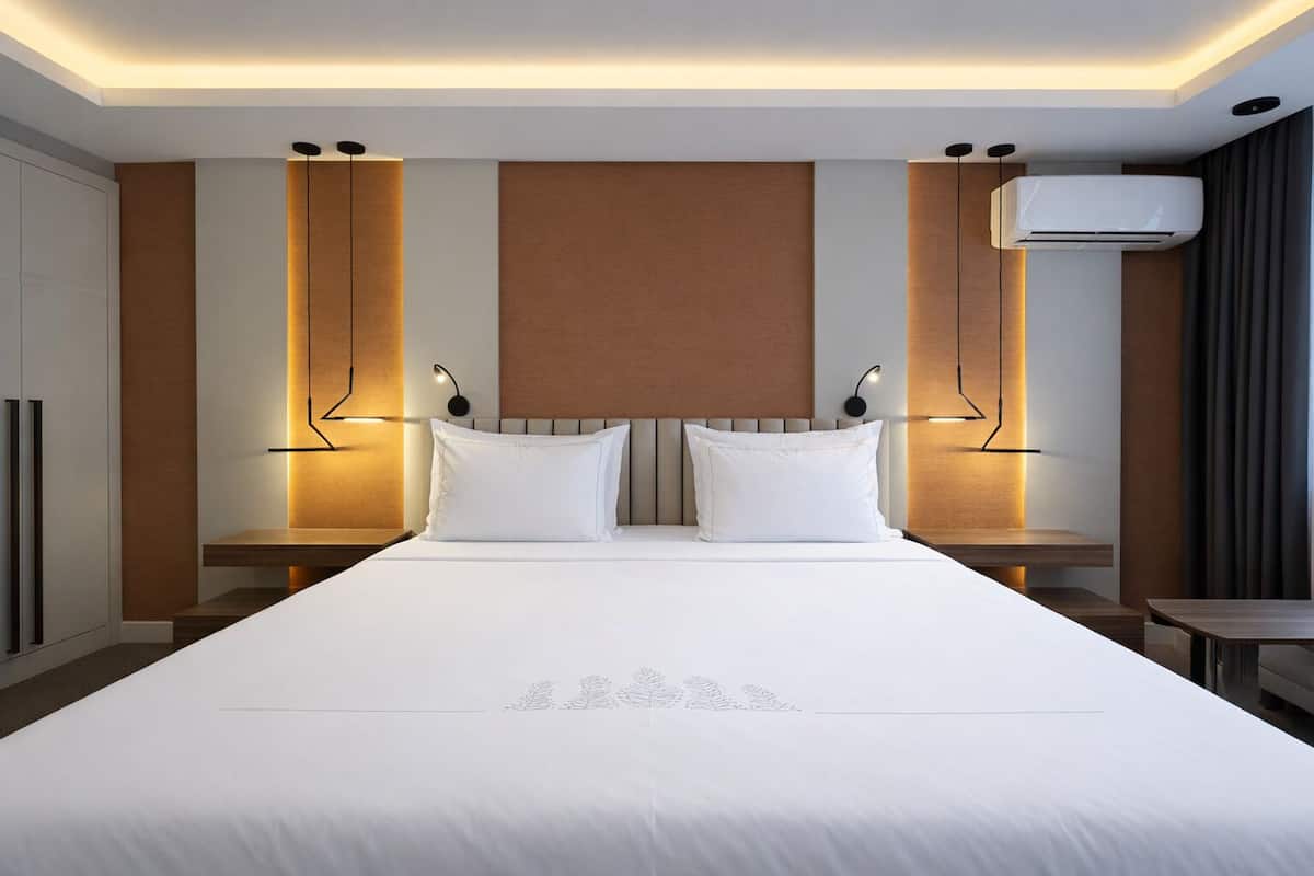 A modern hotel room with a large bed, two bedside tables, a wall-mounted air conditioner, and a ceiling light fixture.