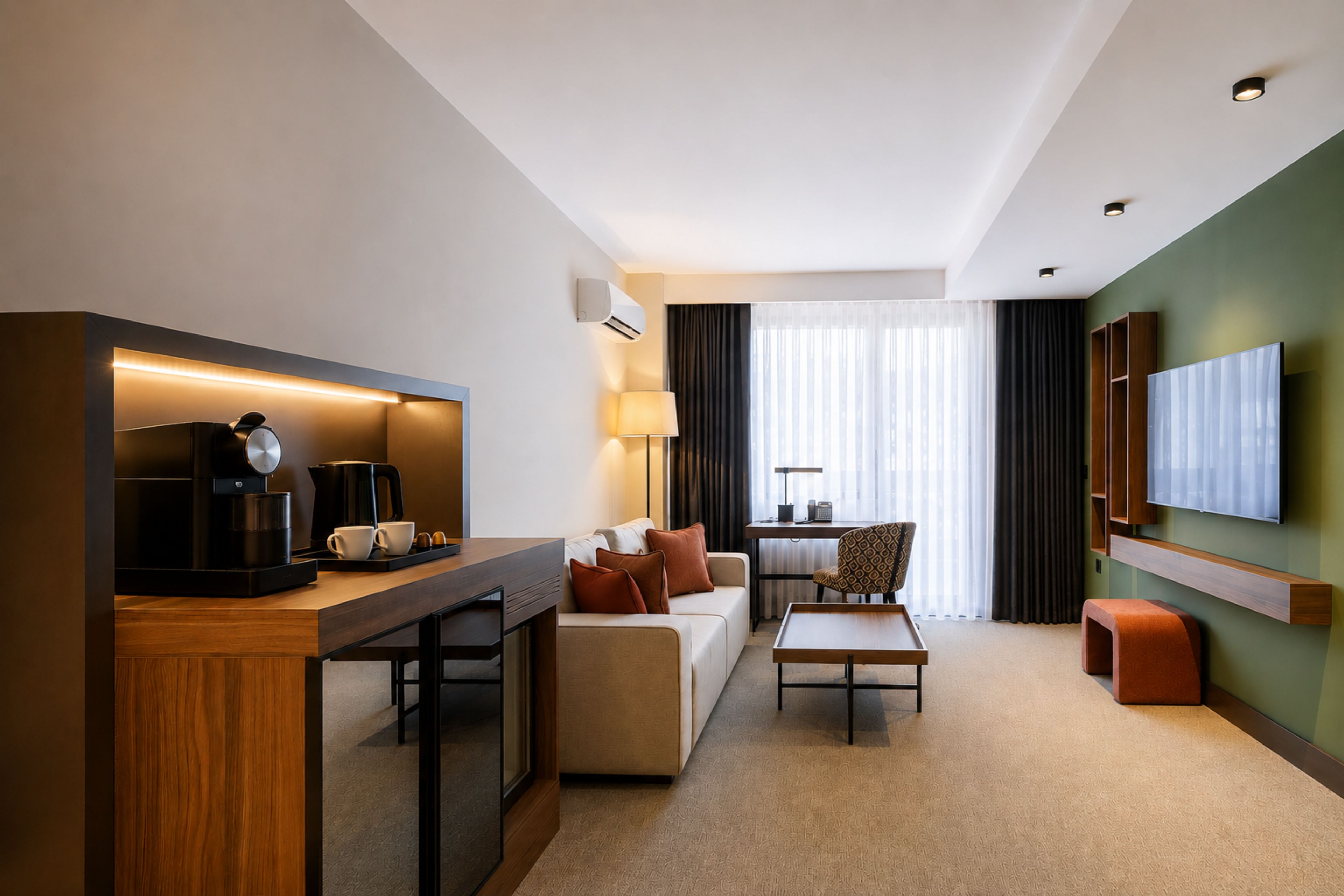 A modern hotel room with a kitchenette, a sofa, a coffee table, a TV, and a large window with curtains.