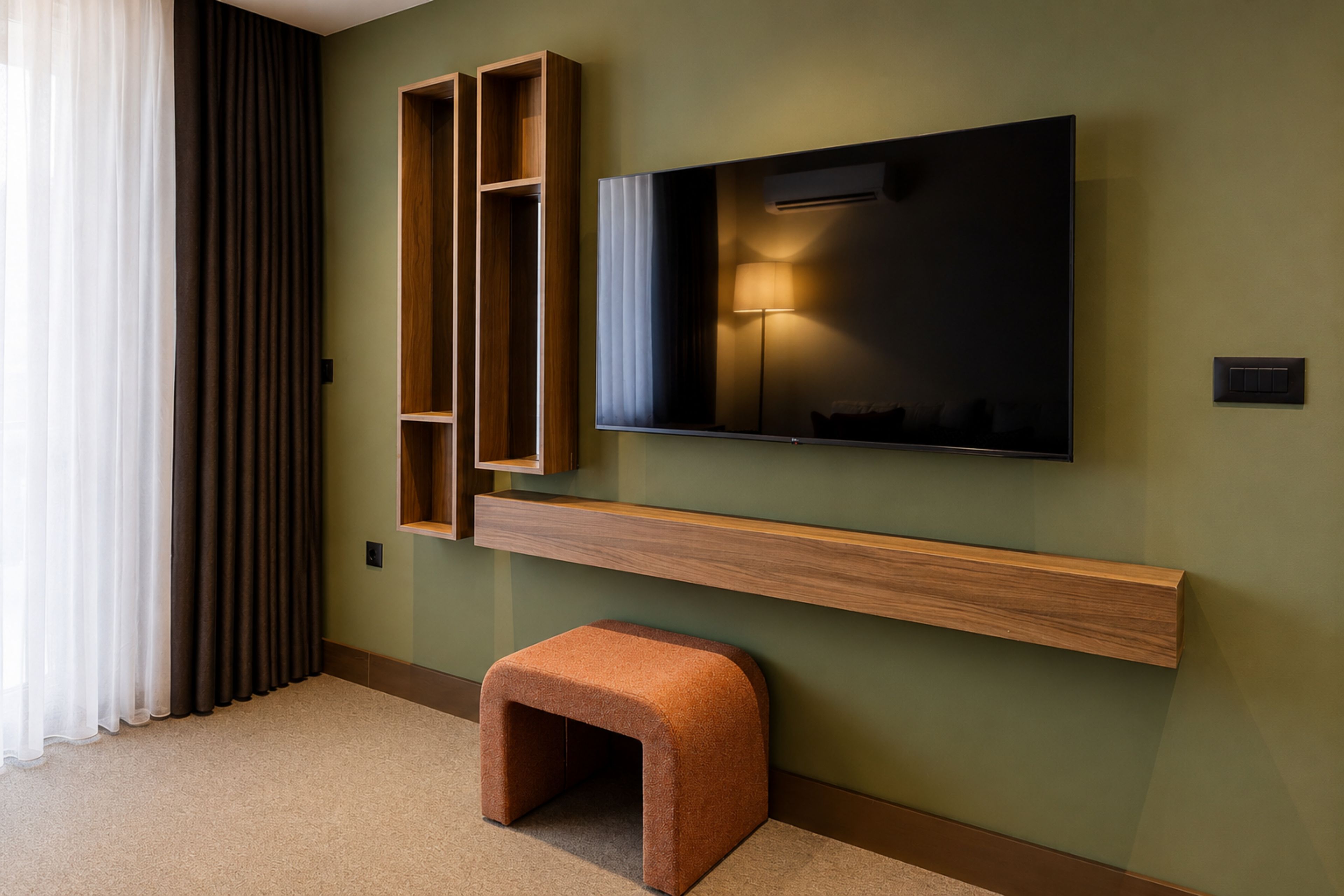Una sala de estar moderna con un televisor de pantalla plana montado en un estante de madera, un taburete pequeño y una cortina.