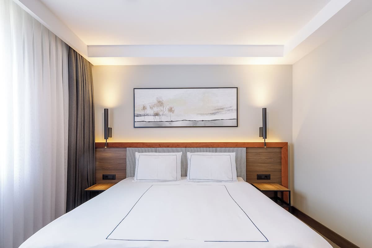 A modern hotel room with a large bed, two bedside tables, a wooden headboard, and a framed artwork above the bed.